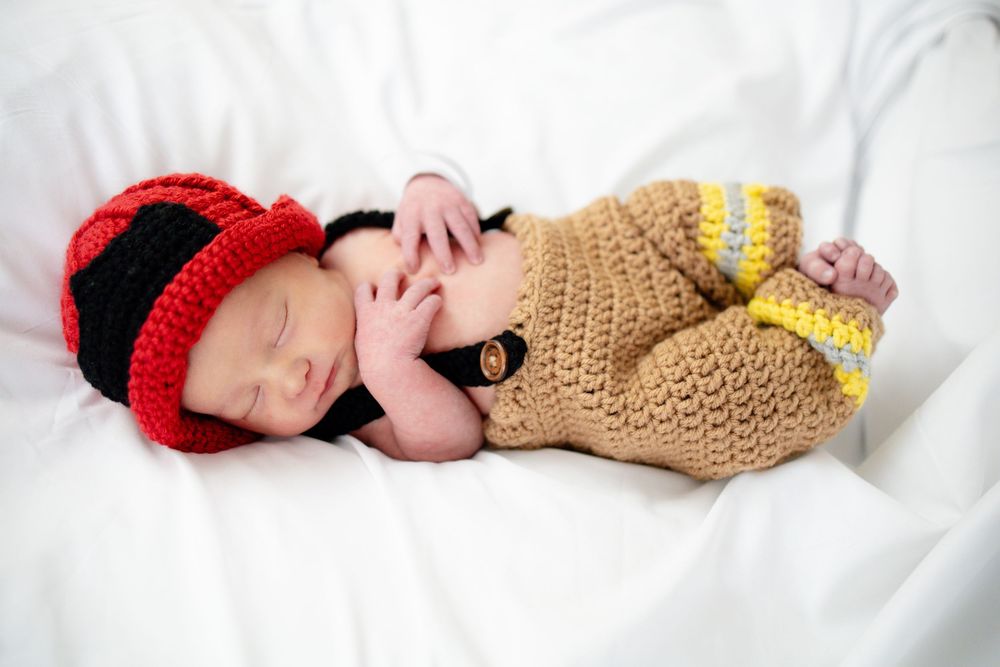 Newborn baby sleeping in crochet firefighter costume with red helmet hat and tan turnout gear outfit.