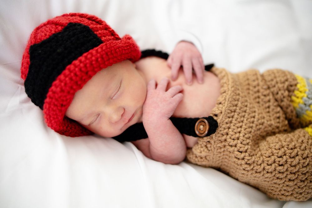Sleeping newborn baby dressed in a crocheted firefighter costume with red and black helmet and tan pants.