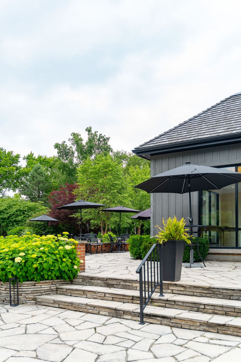 Elegant stone patio with black umbrellas, lush greenery, and modern dark-sided building entrance on a cloudy day.