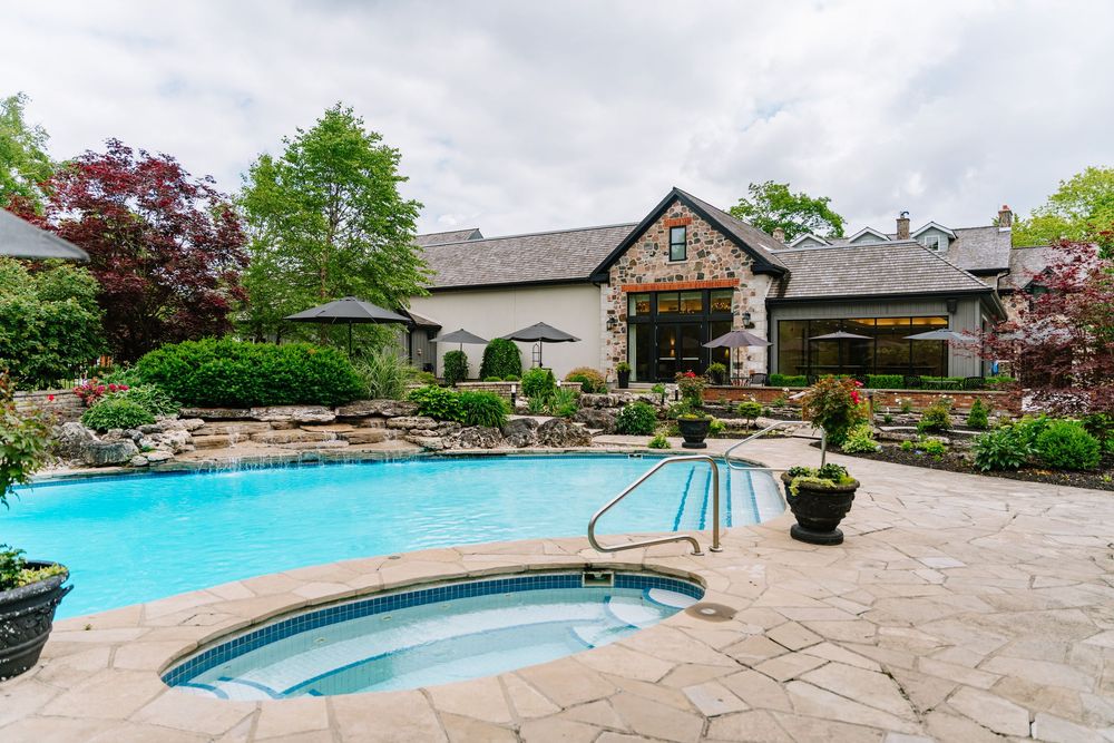 Luxury backyard with a freeform pool, hot tub, stone patio, and elegant stone house surrounded by lush greenery.