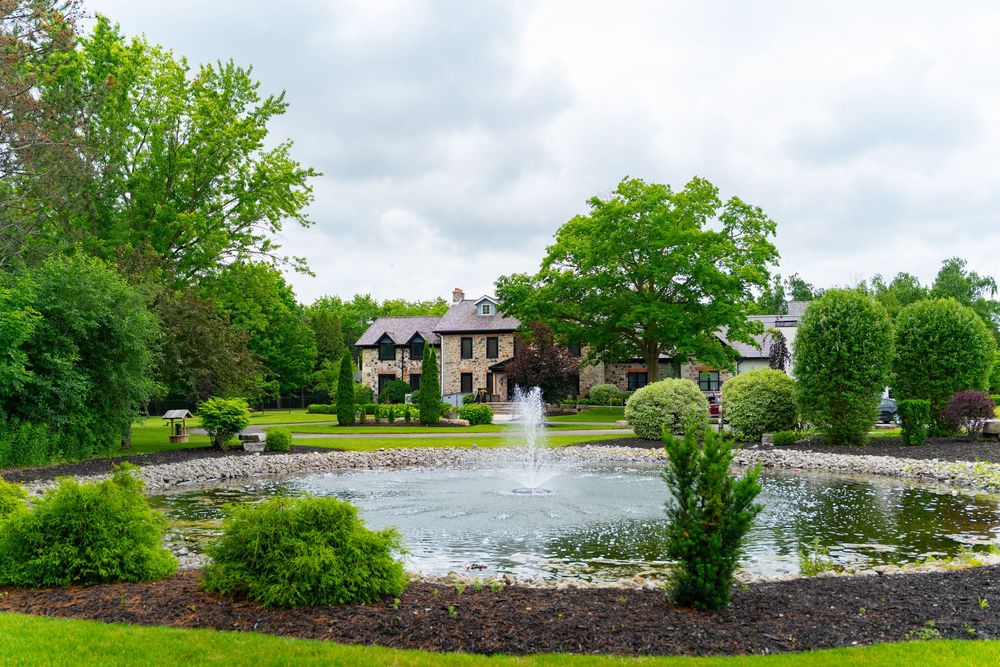 Elegant stone manor house with lush gardens and a decorative pond featuring a fountain on a cloudy day.