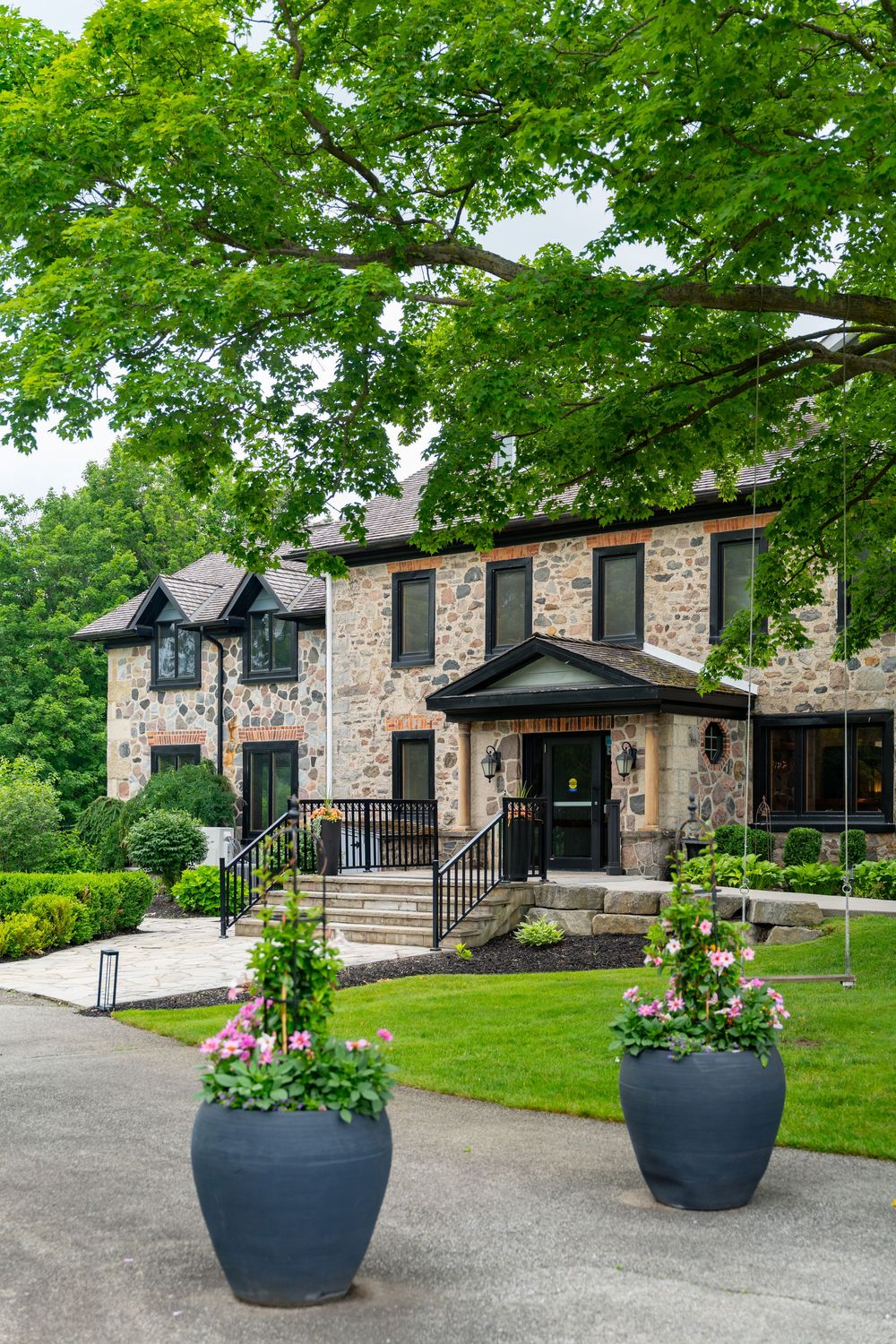 Elegant stone manor house with lush green trees, manicured lawn, and decorative flower planters at the entrance.