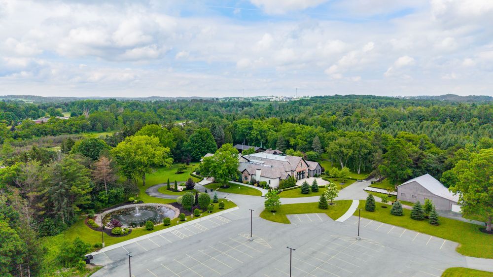 Aerial view of a large estate home surrounded by lush green trees with a circular driveway and expansive parking area.