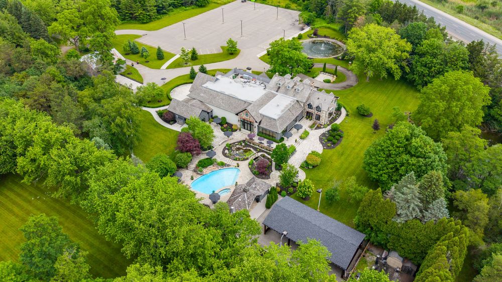 Aerial view of a luxury estate with a pool, manicured gardens, circular driveway, and lush green trees surrounding the property.
