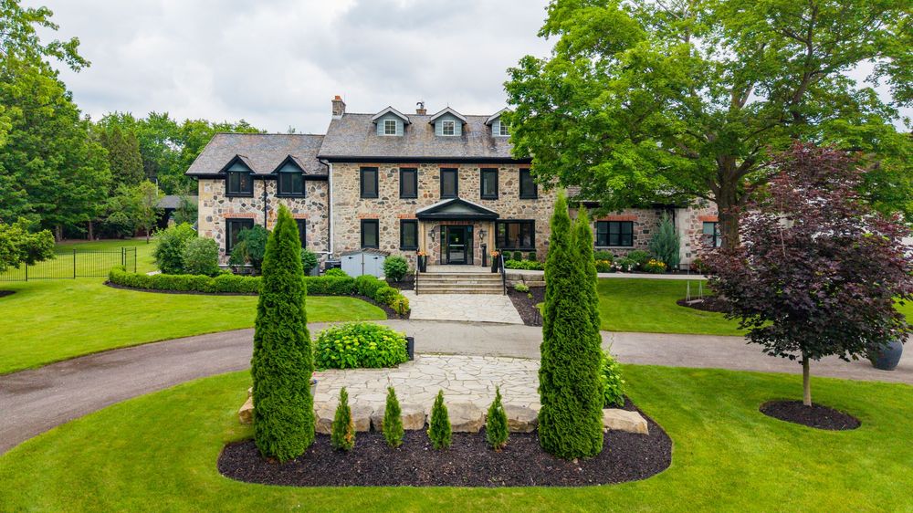 Elegant stone mansion with manicured lawn, tall evergreen trees, and circular driveway surrounded by lush greenery.