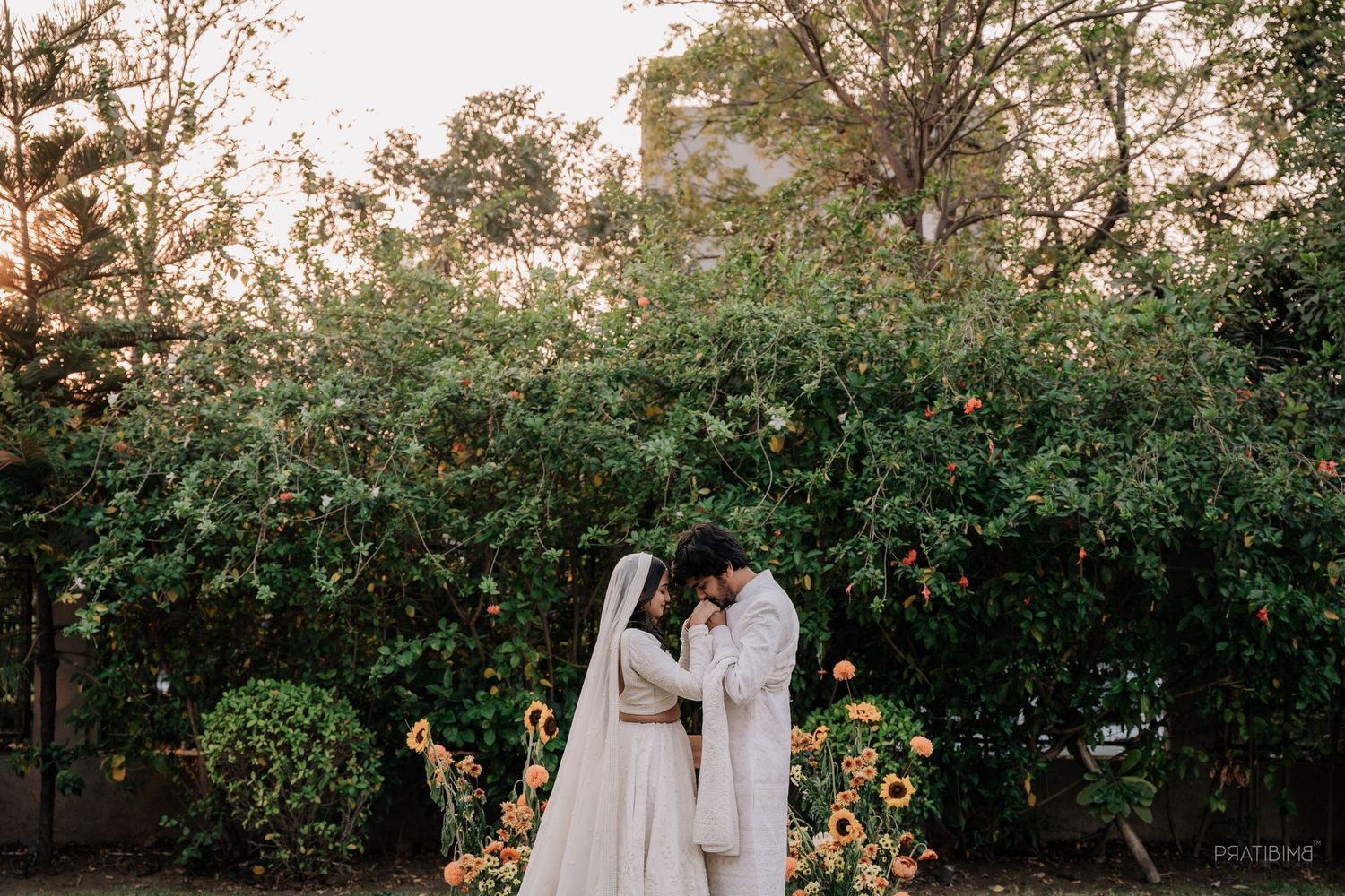 Couple in white wedding attire sharing an intimate moment surrounded by lush greenery and sunflowers in a garden.