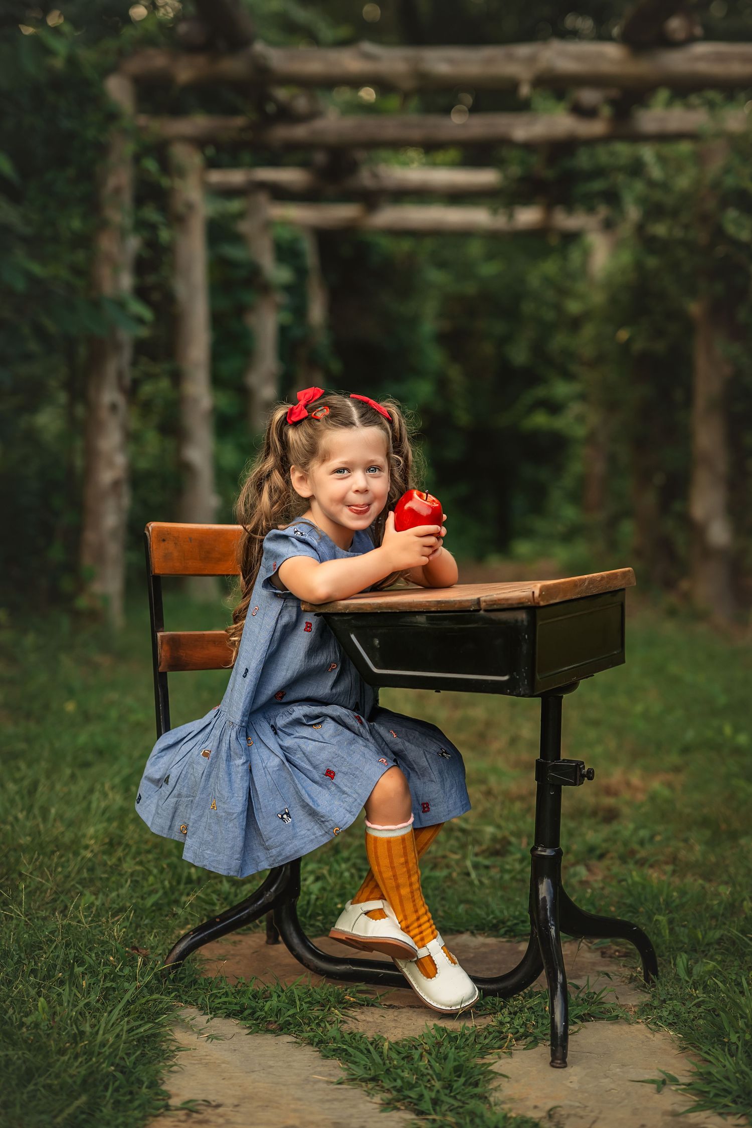 Young girl in blue dress sitting at vintage school desk outdoors, smiling and holding a red apple.
