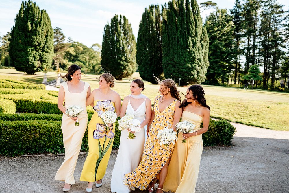 Bridesmaids in colorful floral dresses and pale gowns walk together laughing on a manicured garden lawn path.