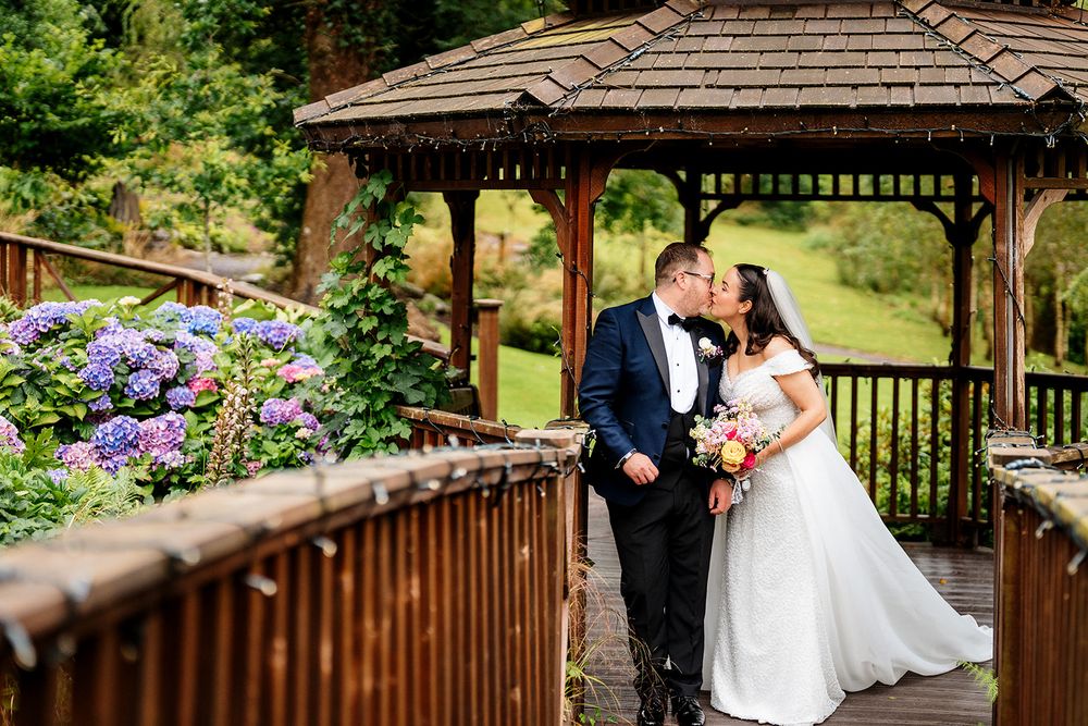 A couple embraces in a rustic wooden gazebo surrounded by vibrant purple hydrangeas in a garden setting.