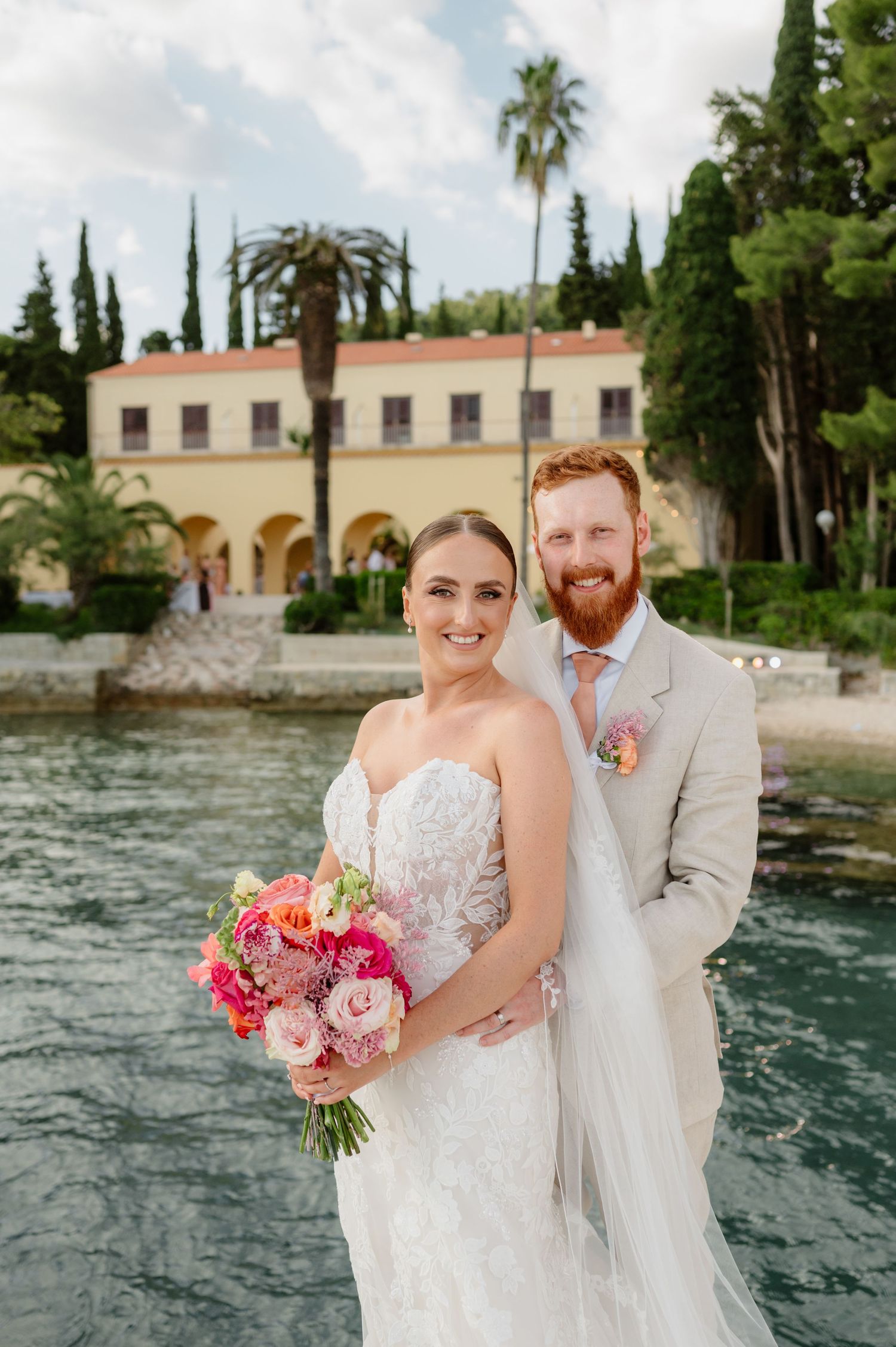 Bride and groom pose by waterfront villa, she holds pink bouquet in lace gown, he wears tan suit with pink tie.