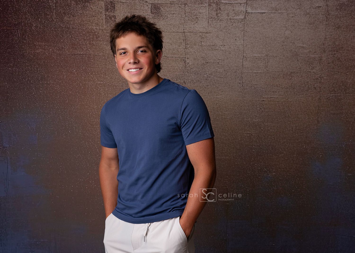 Young man smiling in blue t-shirt and white pants poses confidently against a dark textured studio backdrop.