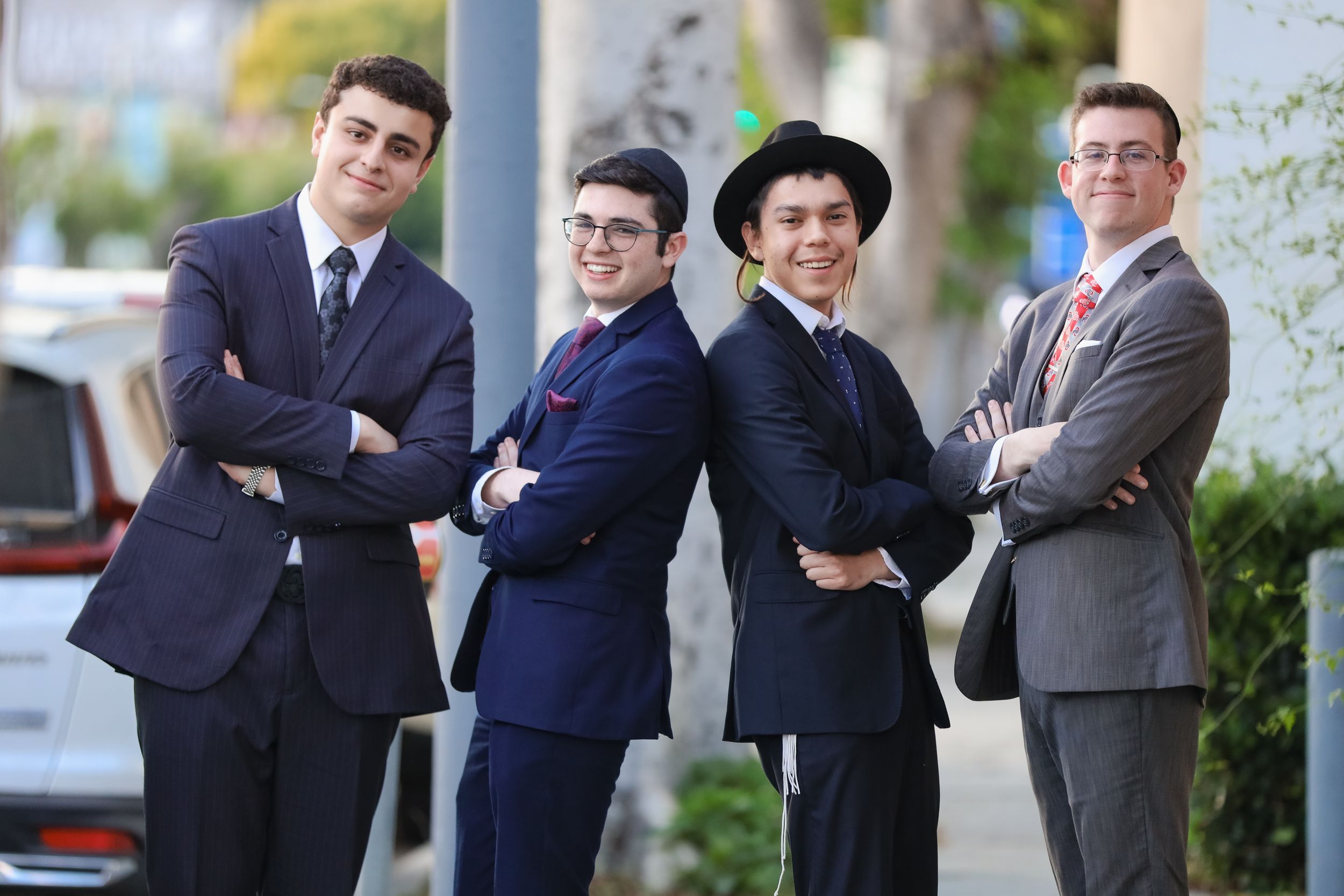 Four young men in suits posing with arms crossed on a sunny street.