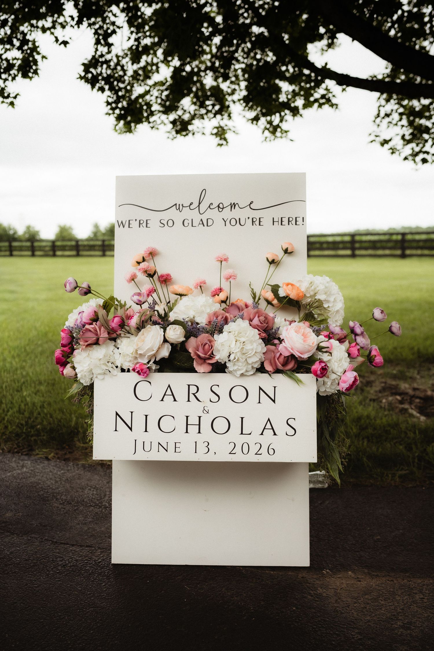 Elegant wedding welcome sign for Carson and Nicholas on June 13, 2026, adorned with pink and white floral arrangements outdoors.