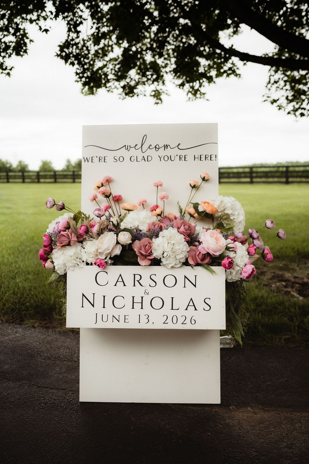 Elegant wedding welcome sign for Carson and Nicholas on June 13, 2026, adorned with pink and white floral arrangements outdoors.