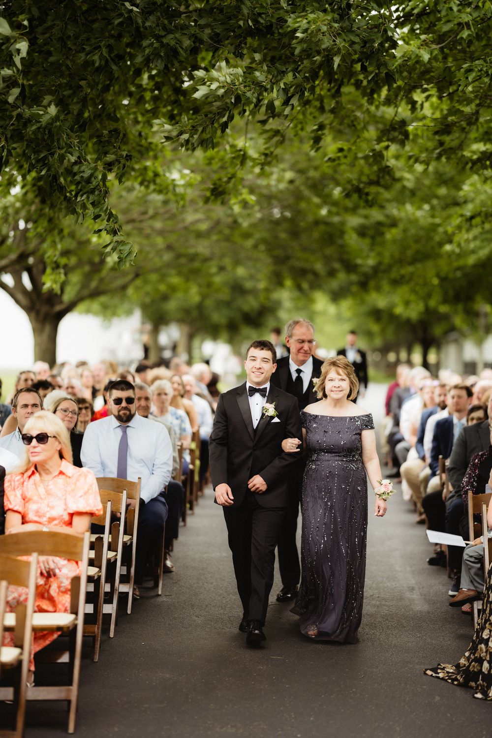 Groom walks down outdoor wedding aisle with mother in sparkly gown, guests seated under lush green trees.