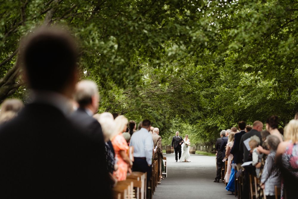 Outdoor wedding ceremony with guests seated on wooden benches lining a tree-canopied aisle as couple walks toward altar.