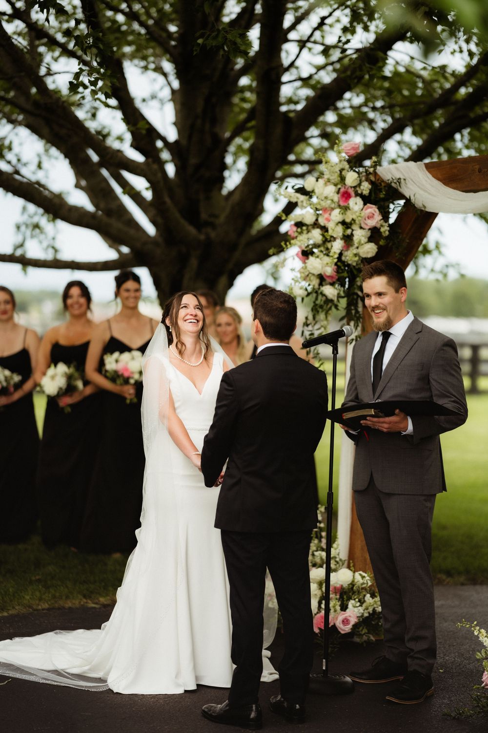Bride laughing joyfully during outdoor wedding ceremony under large tree with bridesmaids in black dresses and floral arch.