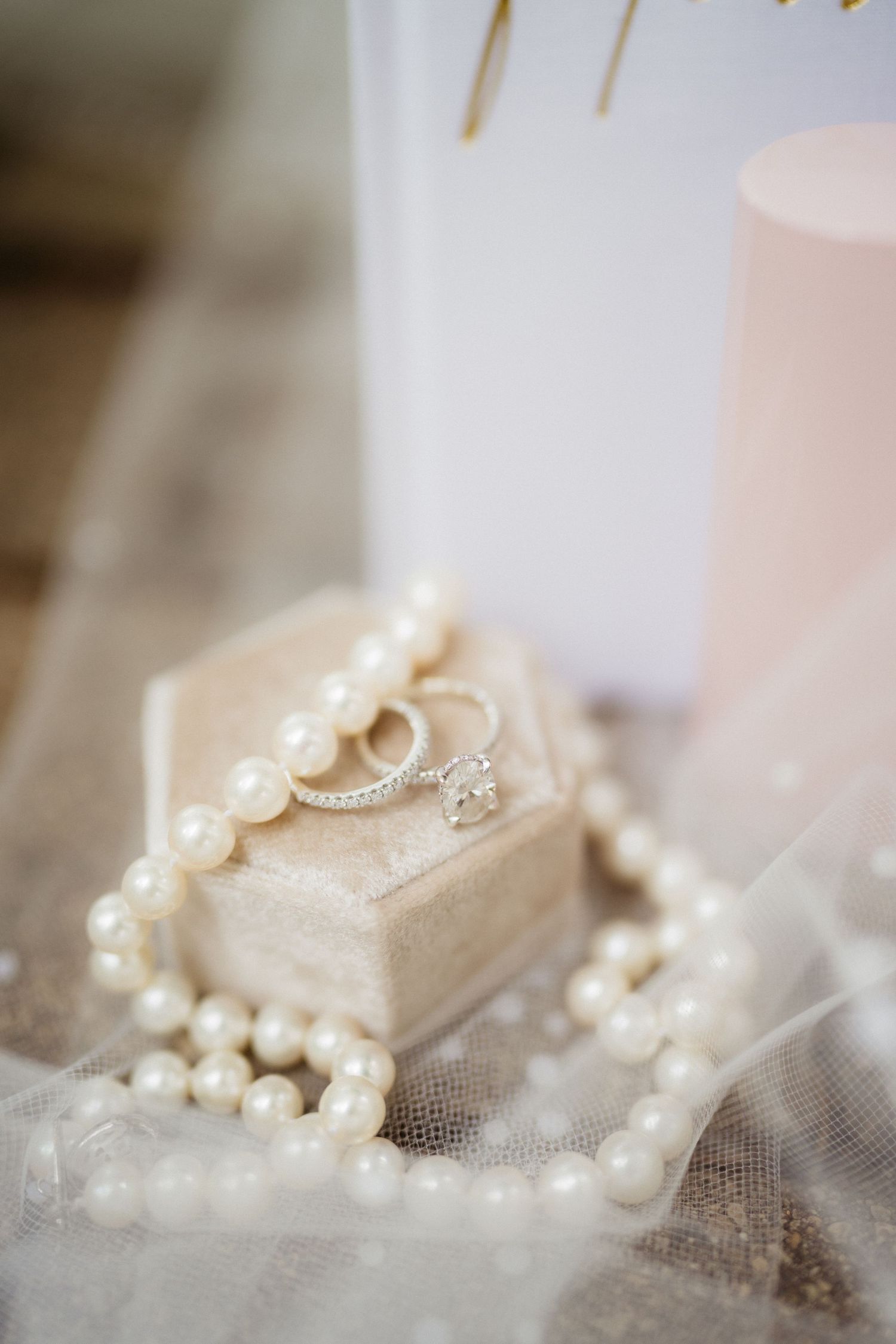Elegant wedding rings on a velvet box surrounded by a pearl necklace and soft candlelight on a lace veil.