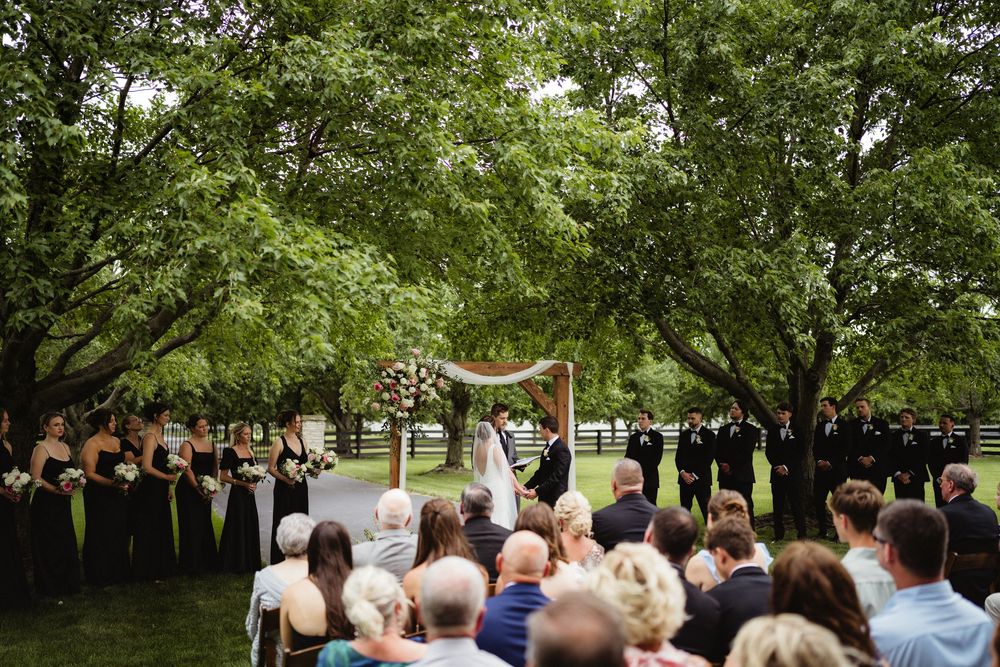 Outdoor wedding ceremony under lush green trees with guests seated, bridal party in dark gowns, and couple at floral arch altar.