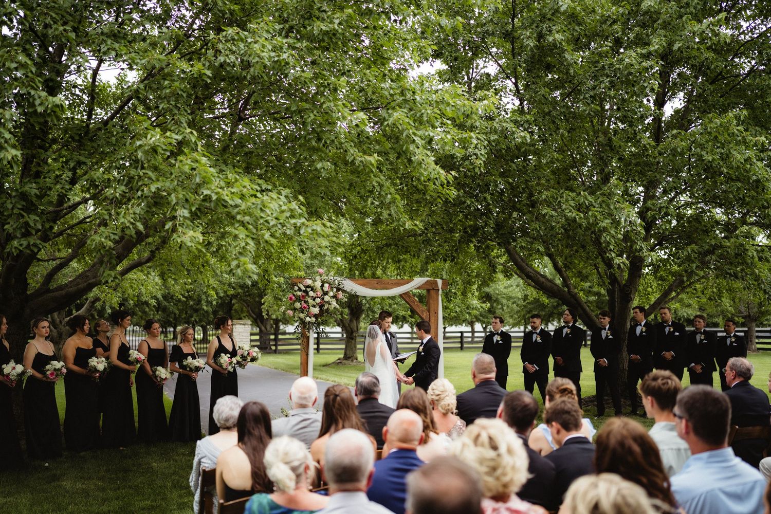 Outdoor wedding ceremony under lush green trees with guests seated, bridal party in dark gowns, and couple at floral arch altar.