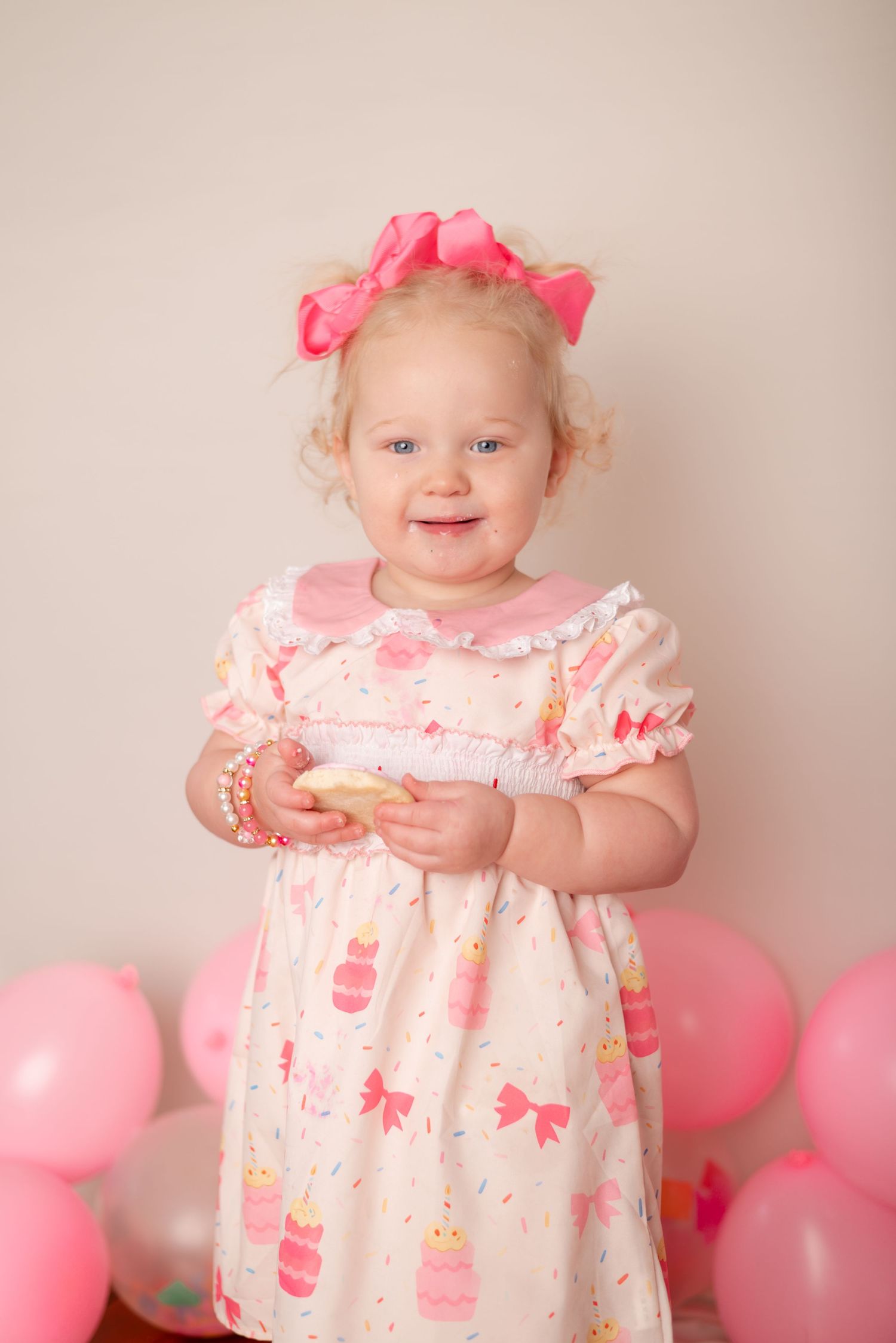 Toddler girl in pink birthday cake dress and bow eating a snack with curly blonde hair and blue eyes.