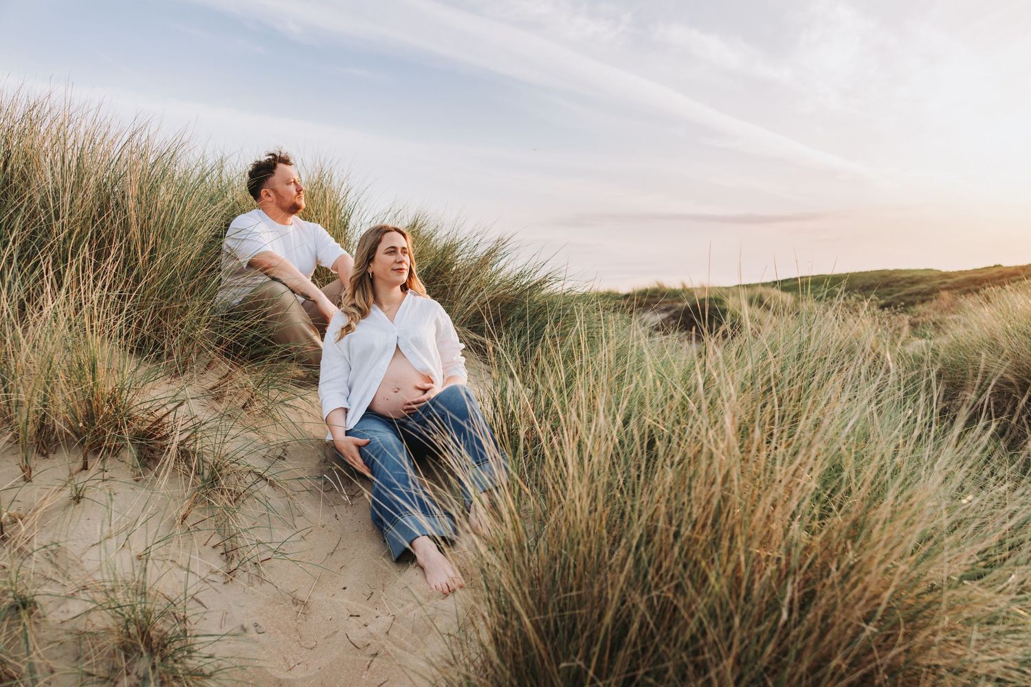 Natural warm edited image of couple sitting on sandy dunes among tall grass at sunset, woman in white shirt touching pregnant belly.