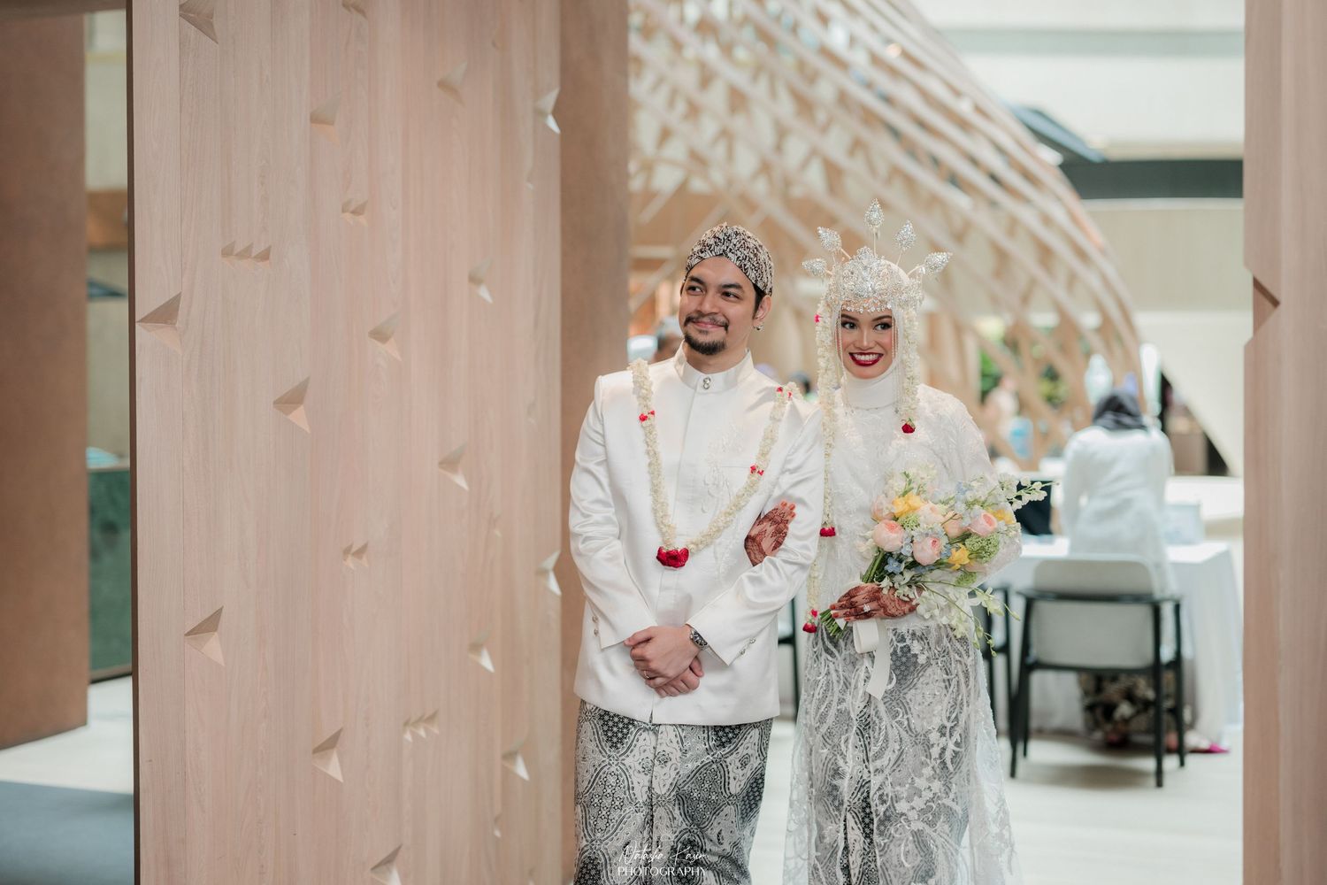Groom in white sherwani and batik sarong with traditional Javanese blangkon hat stands in modern corridor.