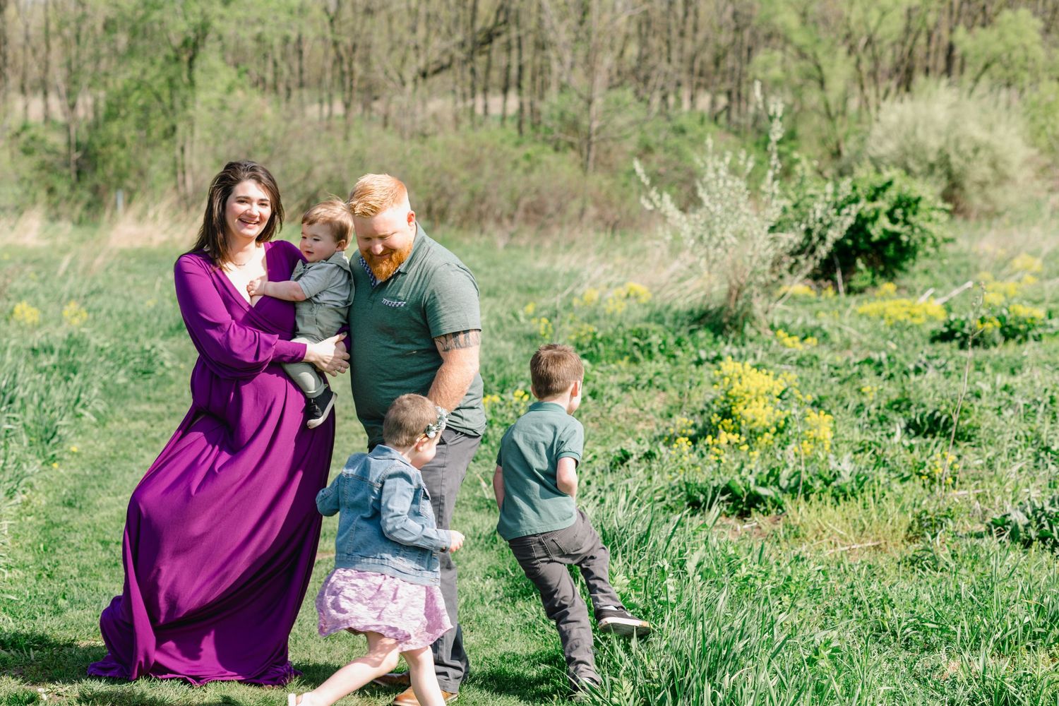 Family of five enjoying a sunny day in a green meadow, mother in purple dress holding baby while children play.