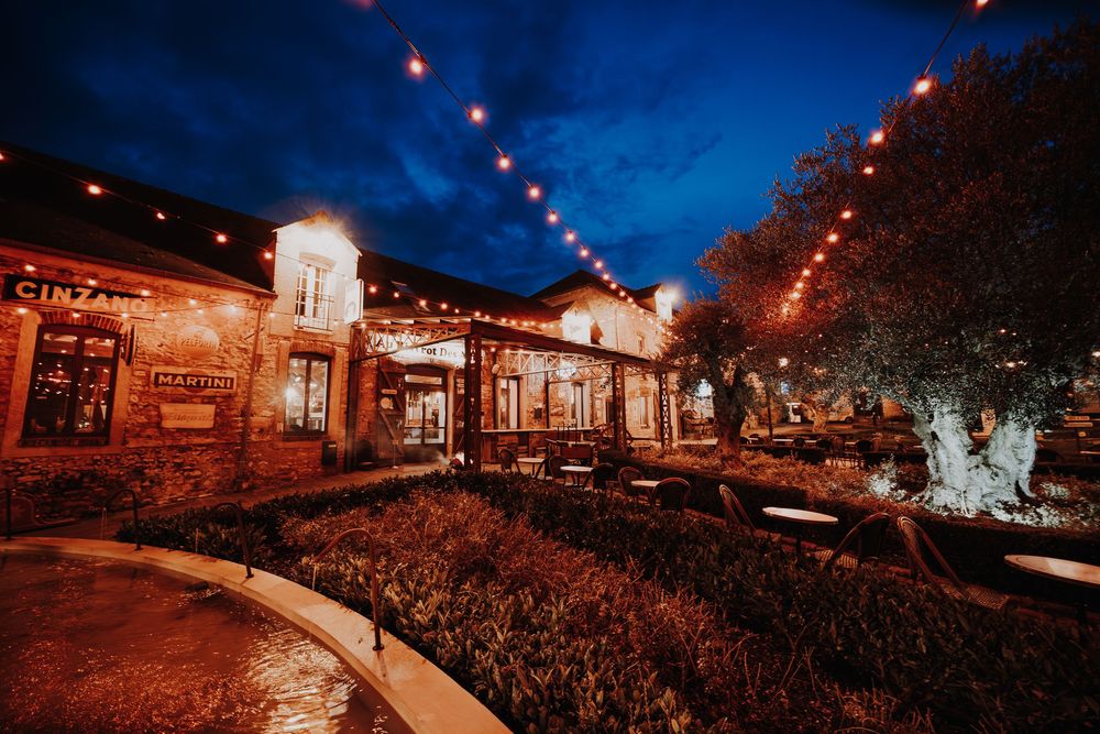 Warm string lights illuminate a charming brick restaurant patio at dusk with lush trees and outdoor seating.