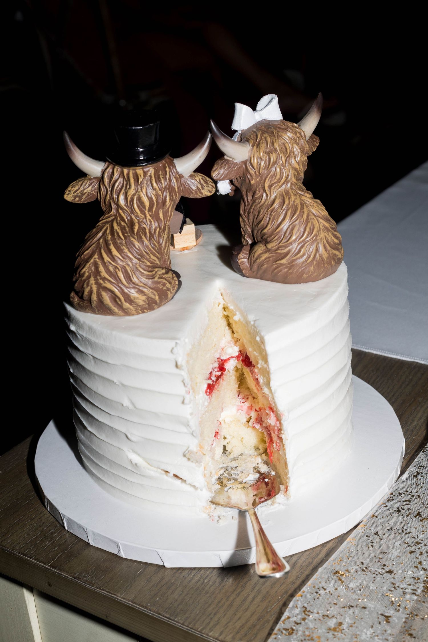 White wedding cake with Highland cow toppers, one slice removed revealing red fruit filling on wooden table.