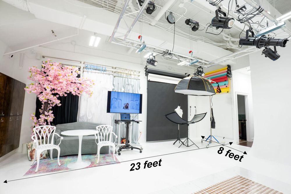 A professional photography studio with pink cherry blossom decor, white chairs, gray backdrops, and studio lighting in a 23x8 ft space.