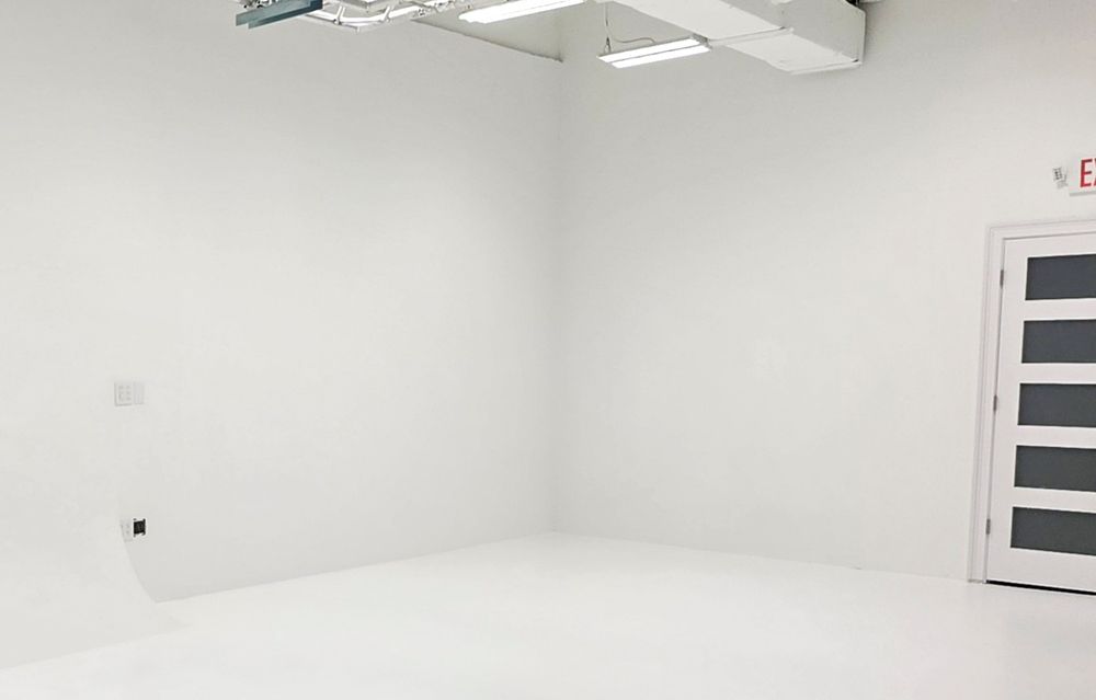 Clean white empty room with bright fluorescent lighting, white walls, epoxy floor, and a white door in the corner.