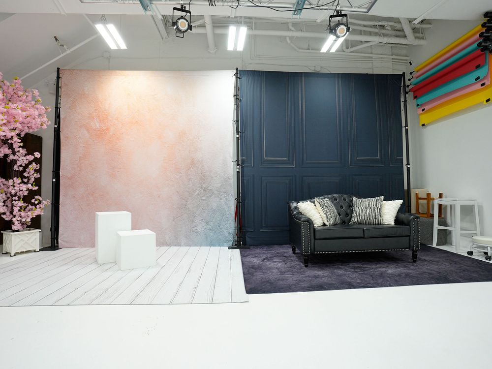 Modern photography studio with pink floral backdrop, navy wall panel, gray sofa, purple rug, and colorful roll backdrops.