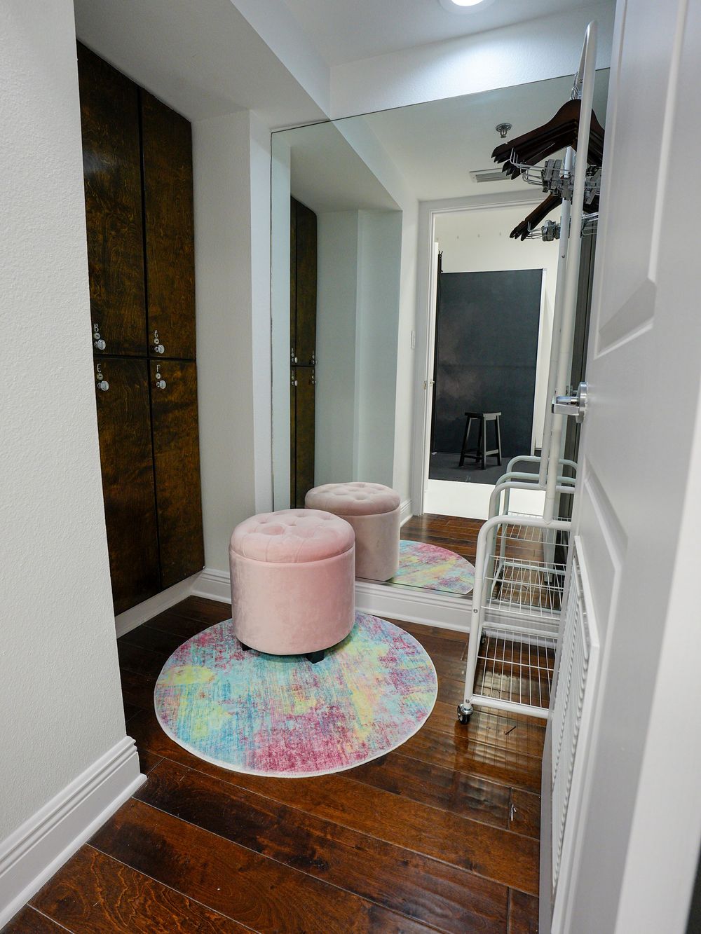 Cozy entryway with pink velvet ottomans on a colorful round rug, large mirror, dark wood floors, and white metal rack.