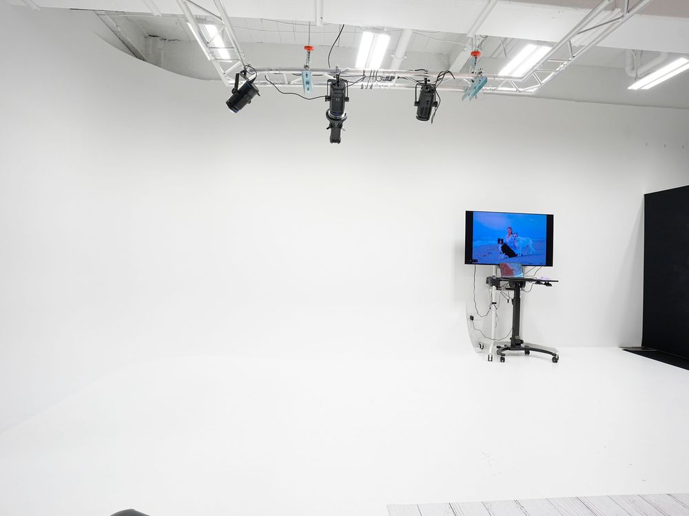 Professional white cyclorama photography studio with overhead lighting rigs and a monitor on a mobile stand.