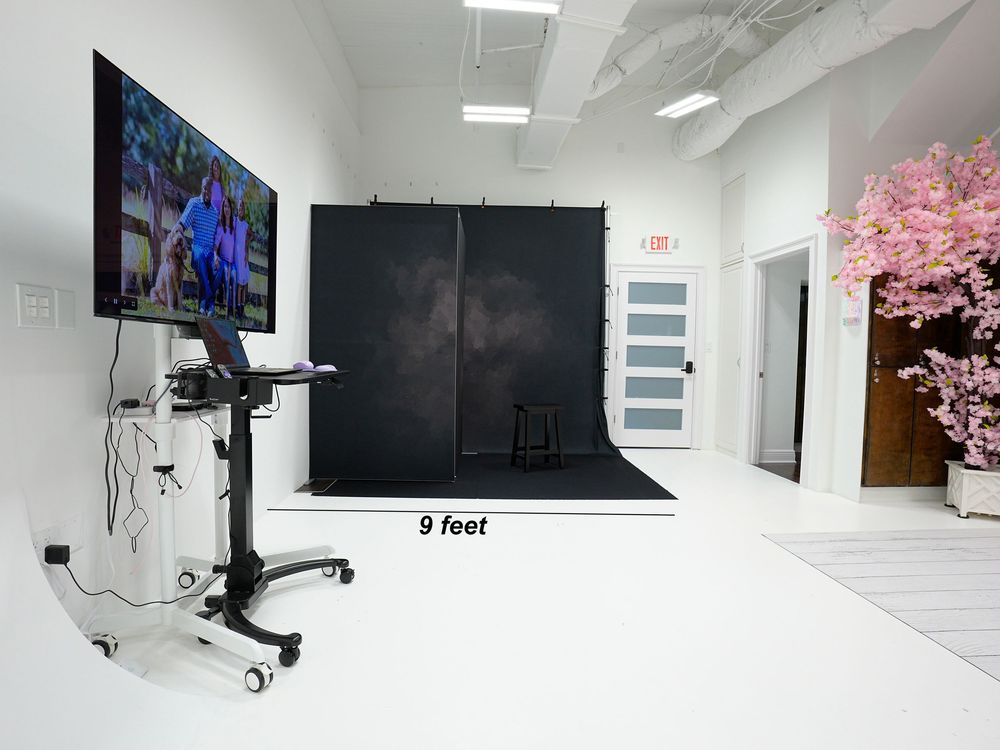 Modern photography studio with black backdrop, TV monitor on stand, pink cherry blossom tree, and white floors spanning 9 feet.