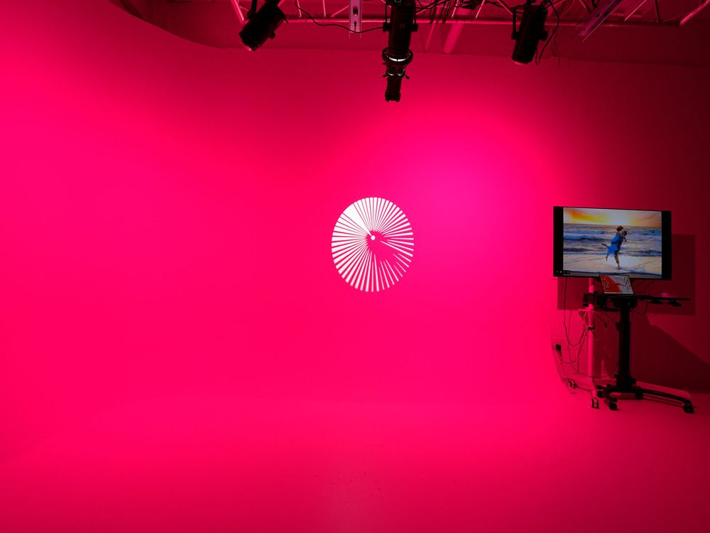 Vibrant pink-lit studio space featuring a white paper umbrella wall art and a monitor displaying a surfer on a beach.
