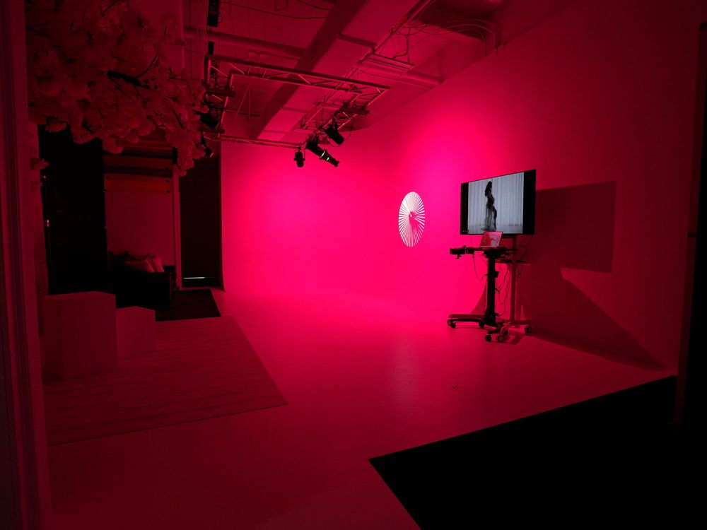 A vibrant pink-lit art installation room with a monitor on a stand and circular display against a glowing magenta wall.