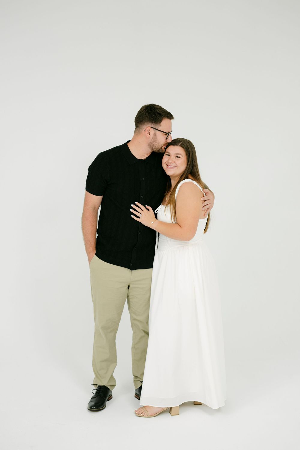 A couple poses together in a studio, man in black shirt kissing woman's head as she smiles in a white dress.