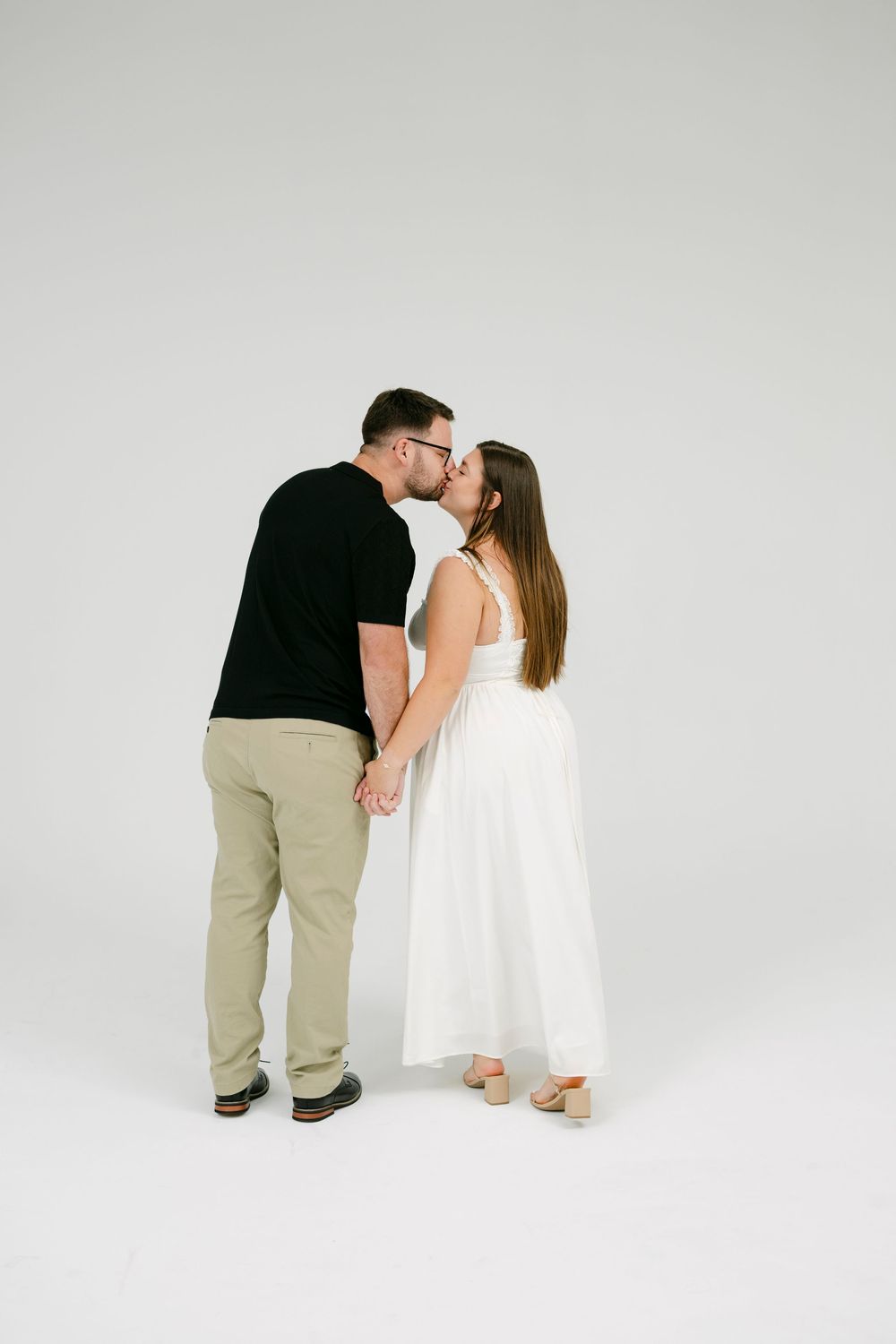 A couple shares a kiss while holding hands, the woman in a white dress and man in black shirt and khaki pants.