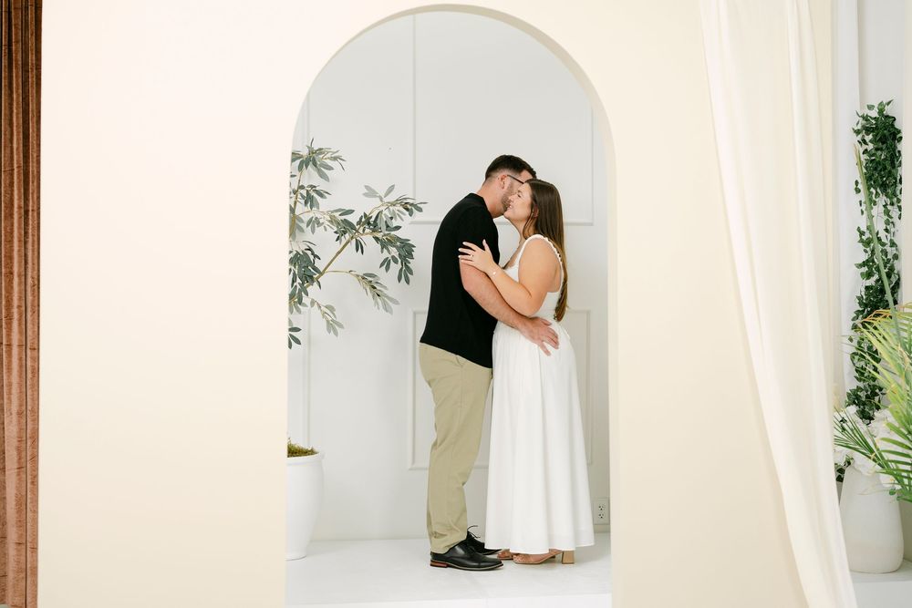 Couple sharing a romantic kiss under an elegant arched doorway in a bright, modern white interior space.