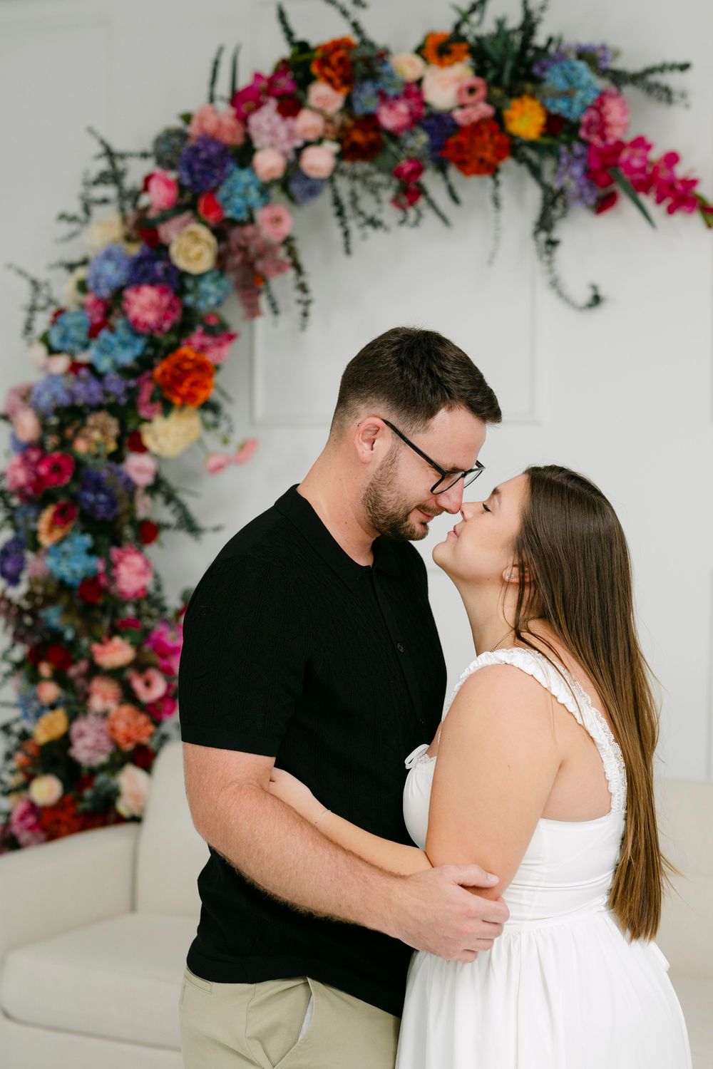 Couple sharing a romantic kiss in front of a colorful floral arch backdrop in a bright white studio.