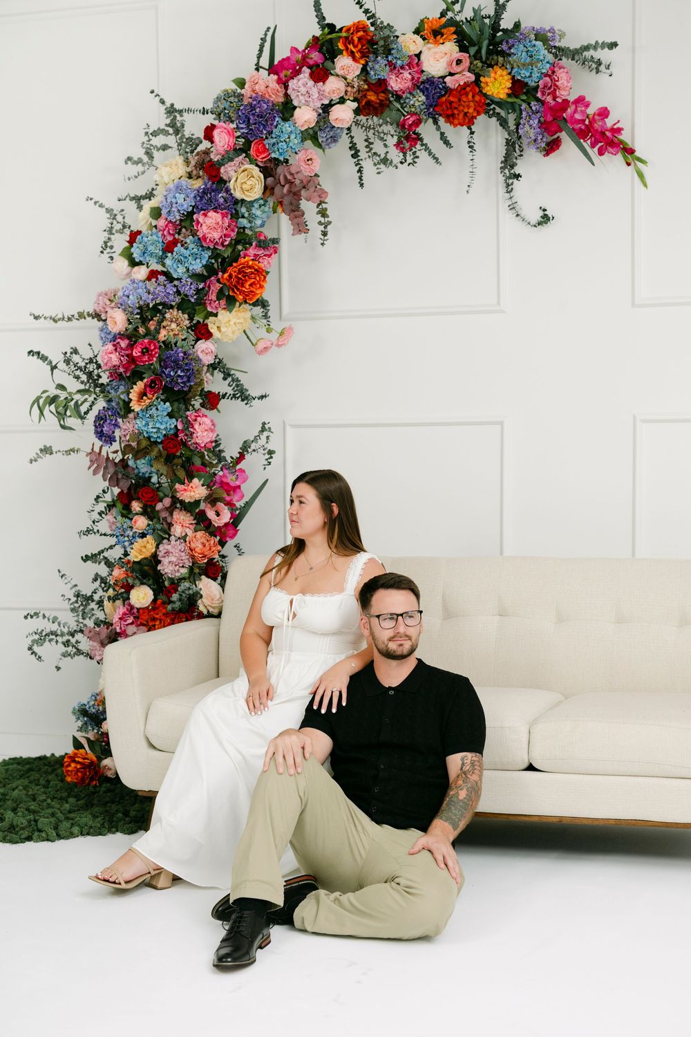 Couple poses by colorful floral arch on white sofa, woman in white dress, man in black shirt, vibrant blooms backdrop.