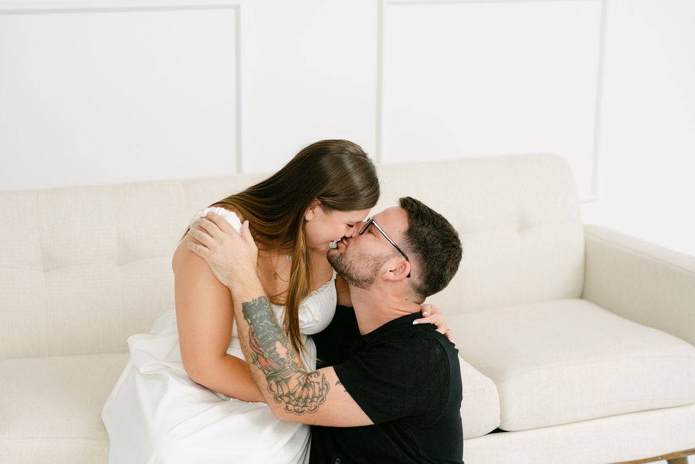 A couple sharing a romantic kiss on a white couch, woman with tattoos embracing a man in a bright white room.