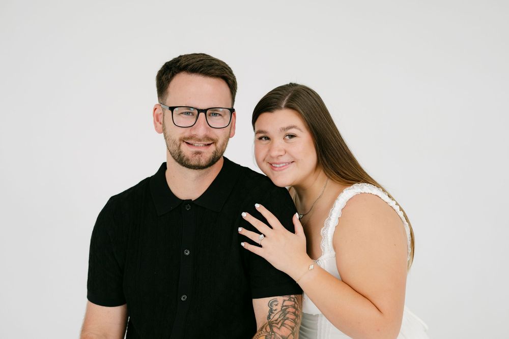 Smiling couple posing together, man in black polo with glasses, woman in white top showing engagement ring.