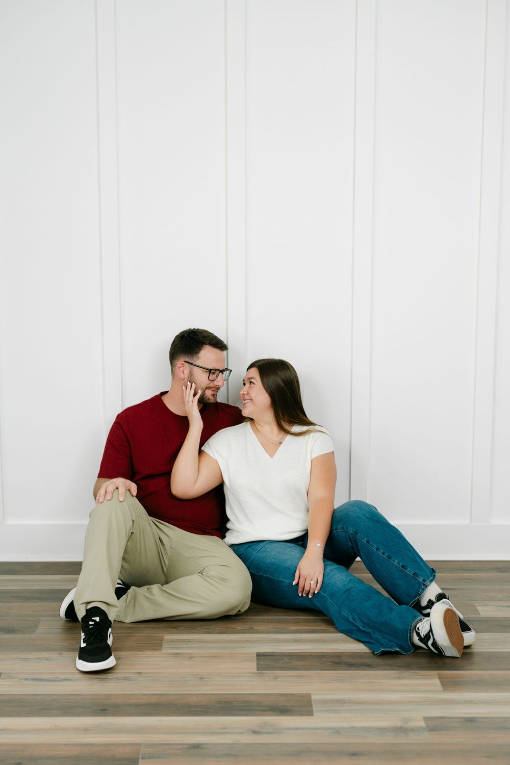 Couple sitting on hardwood floor against white wall, smiling at each other, man in red shirt and glasses, woman in white top.