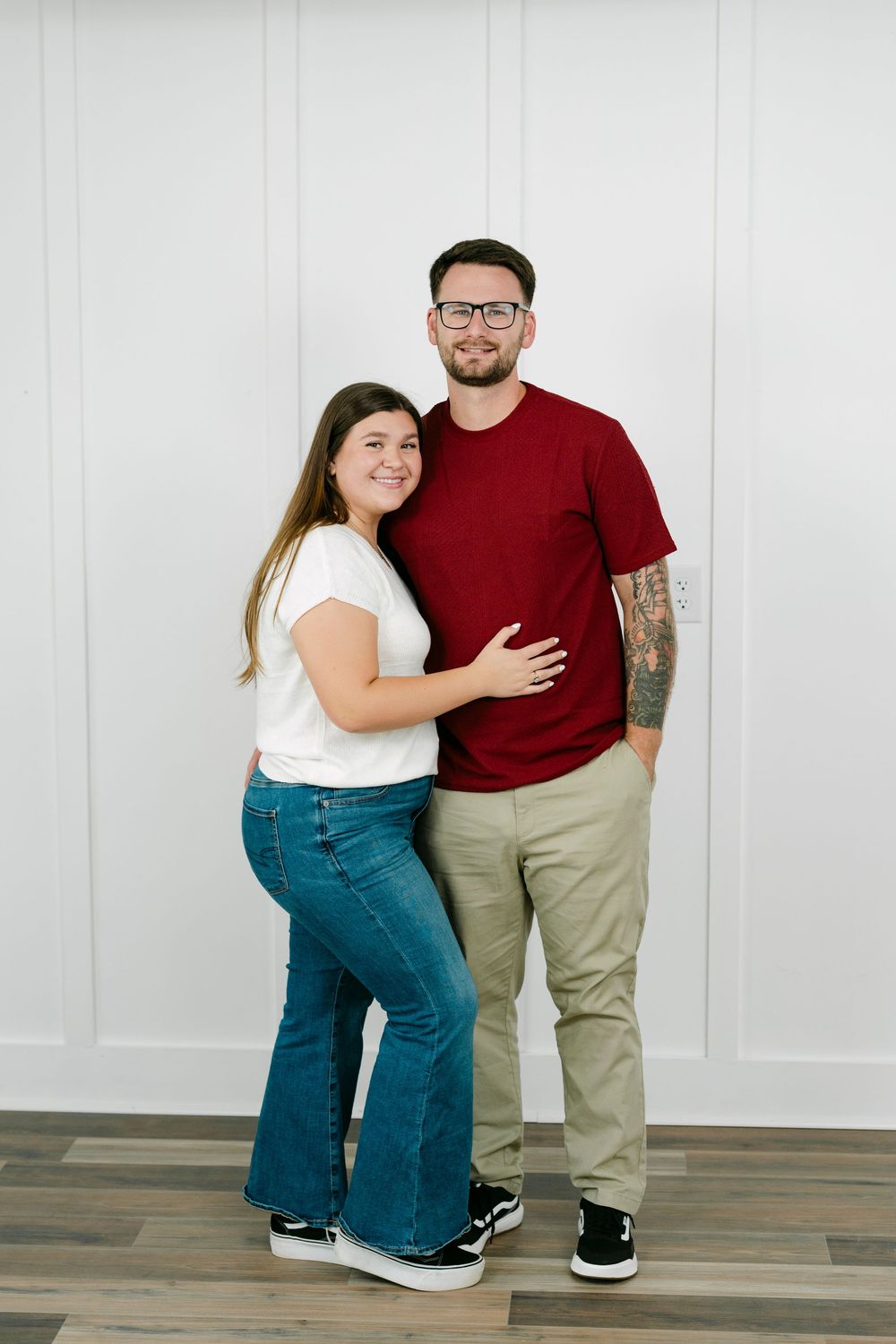 A smiling couple poses together, woman in white tee and flare jeans, man in red shirt and khakis with tattoos.