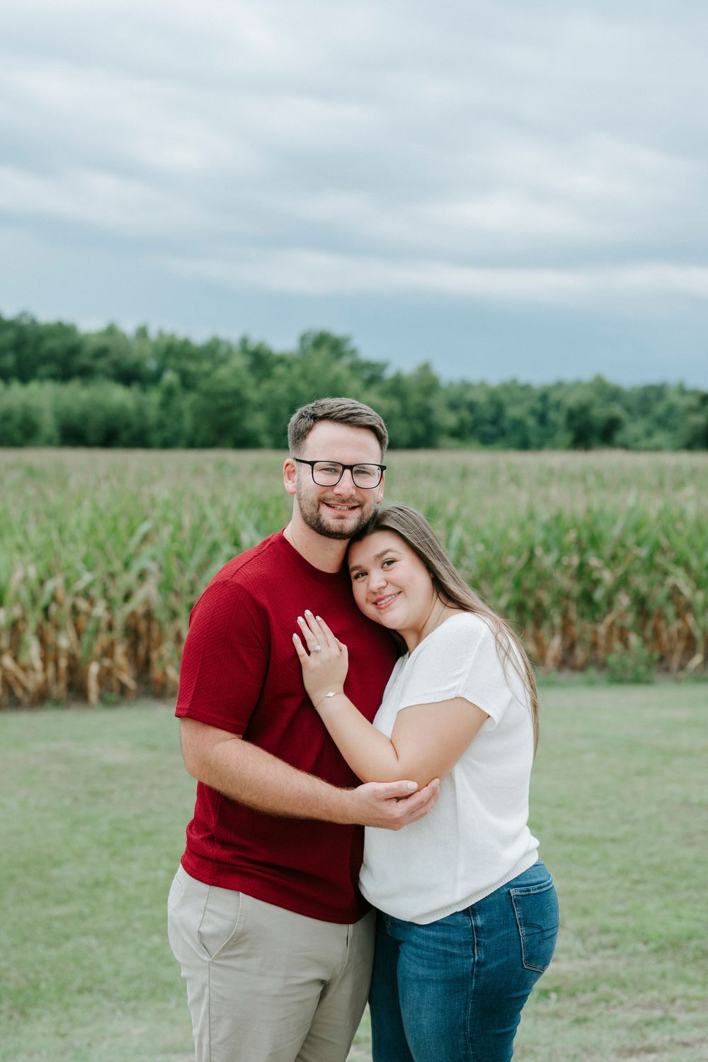 A couple embraces and smiles outdoors near a cornfield, man in red shirt and woman in white top and jeans.