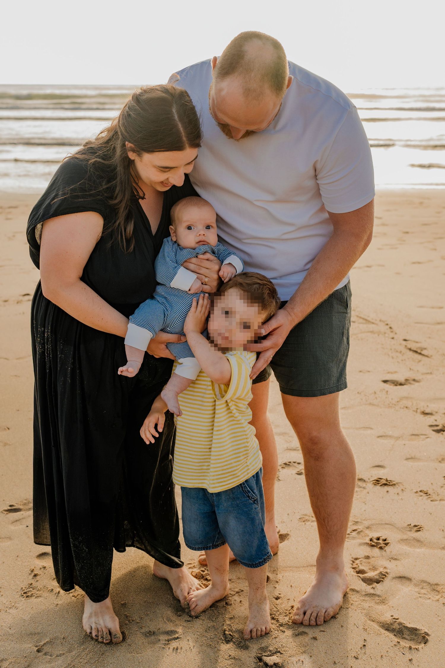 Family of four poses barefoot on sandy beach, parents holding baby and toddler in golden hour light.