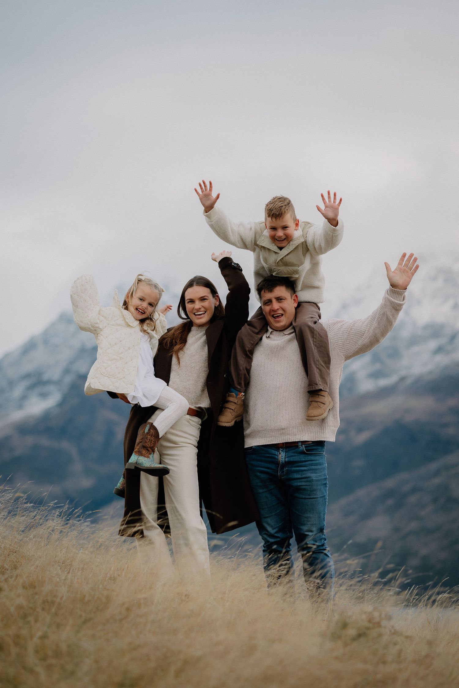 Happy family of four posing with raised hands in a snowy mountain landscape, wearing cozy neutral tones.